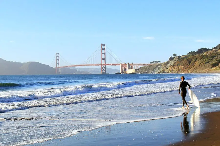 Surfer walking toward water on Baker Beach, bridge on the horizon